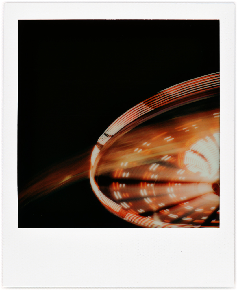 A Polaroid photo of the Flying Circus ride at the 2022 Three Rivers Festival in Fort Wayne, Indiana. The ride is spinning so fast that it is a blur of colored lights against the black night sky.