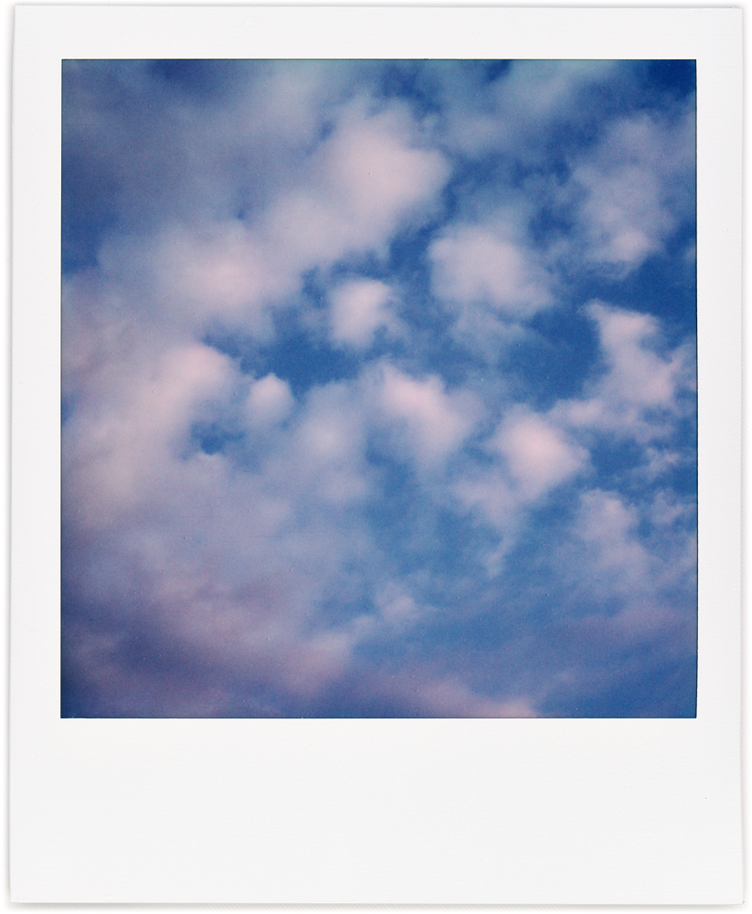 Polaroid abstract photograph of a crescent shaped white cloud formation surrounding a group of small round clouds in the blue morning sky in Fort Wayne, Indiana.