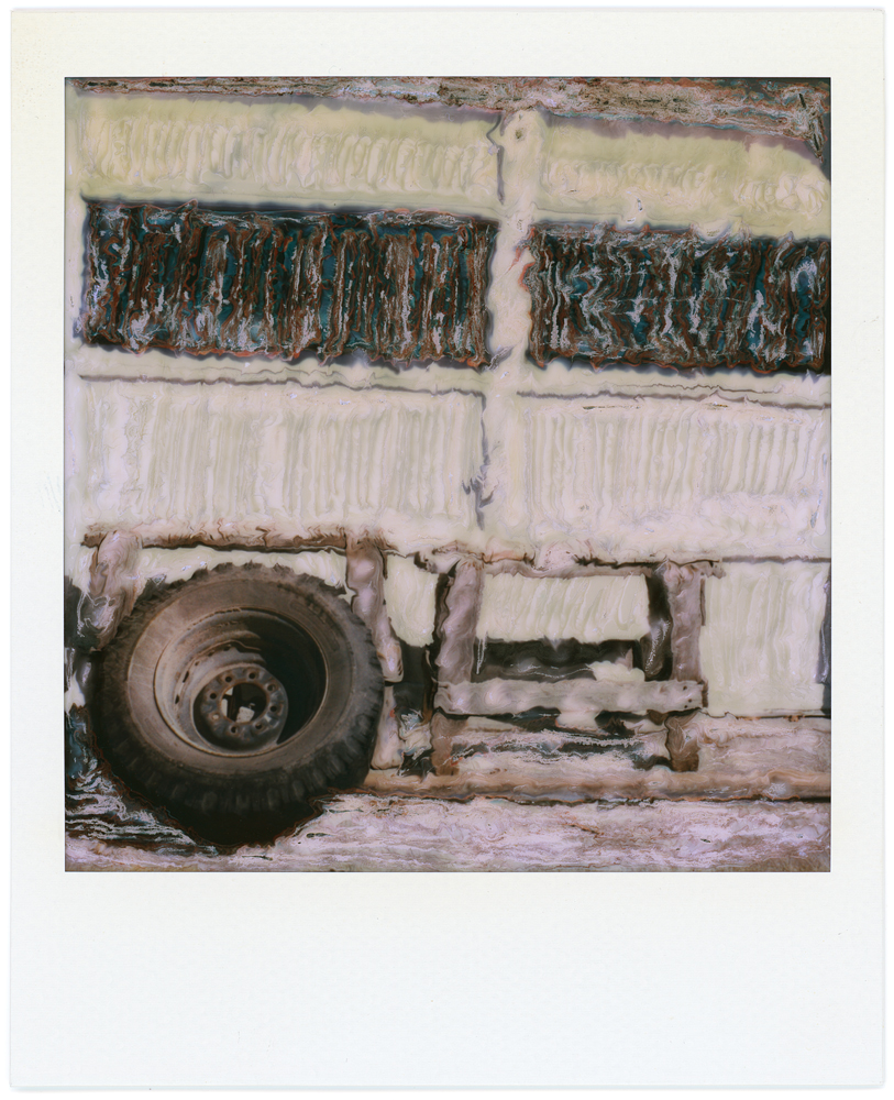 Polaroid SX-70 manipulation photograph of an old truck tire and wheel leaning against a sawhorse in front of a garage door.