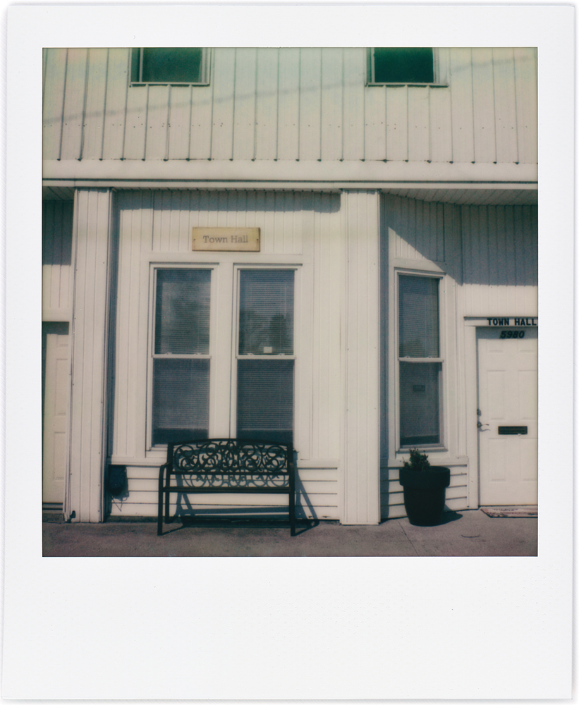 Polaroid snapshot of the town hall in the small town of Uniondale, Indiana. A small white building with a park bench on the sidewalk under the front windows.